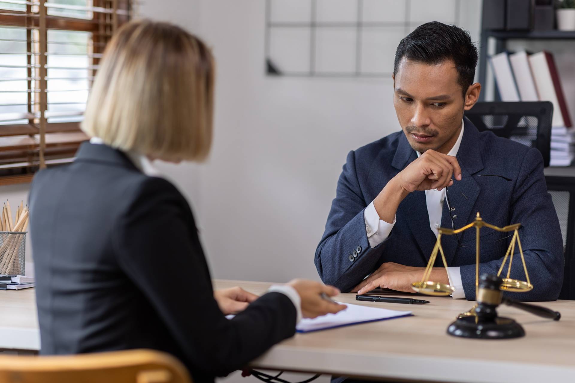 A serious-looking man in a suit sits at a desk with legal scales and a gavel, listening to a woman across from him holding a clipboard